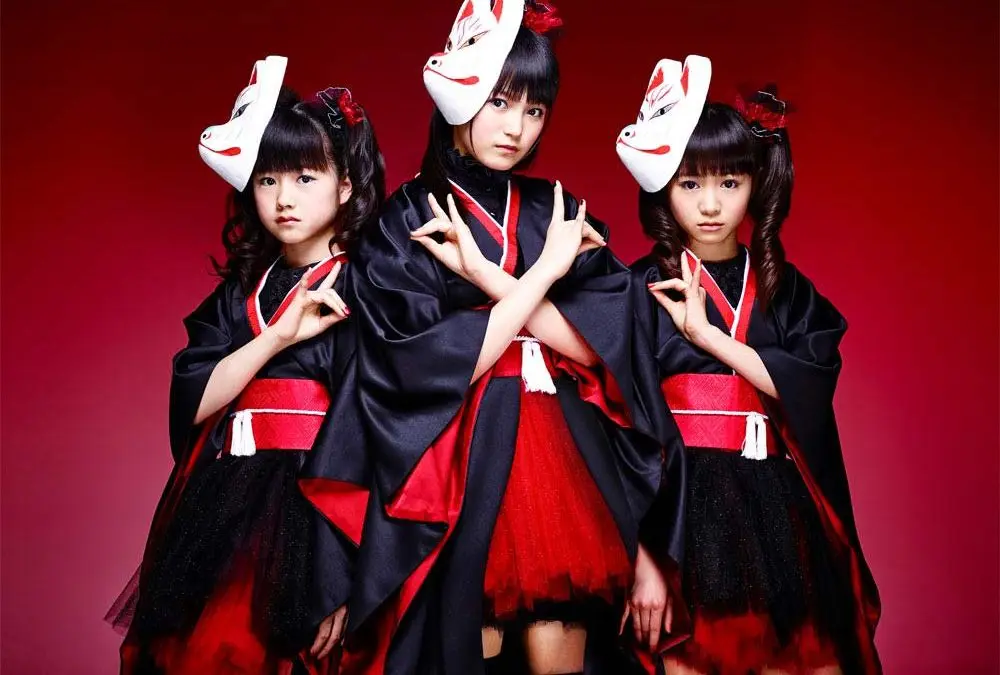 Three young girls in black and red Japanese-inspired outfits with fox masks on their heads pose against a red background…