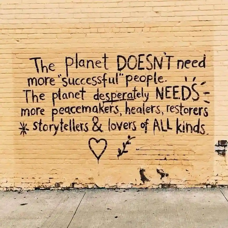 A yellow brick wall has black graffiti text that reads: 'The planet DOESN'T need more "successful" people. The planet…
