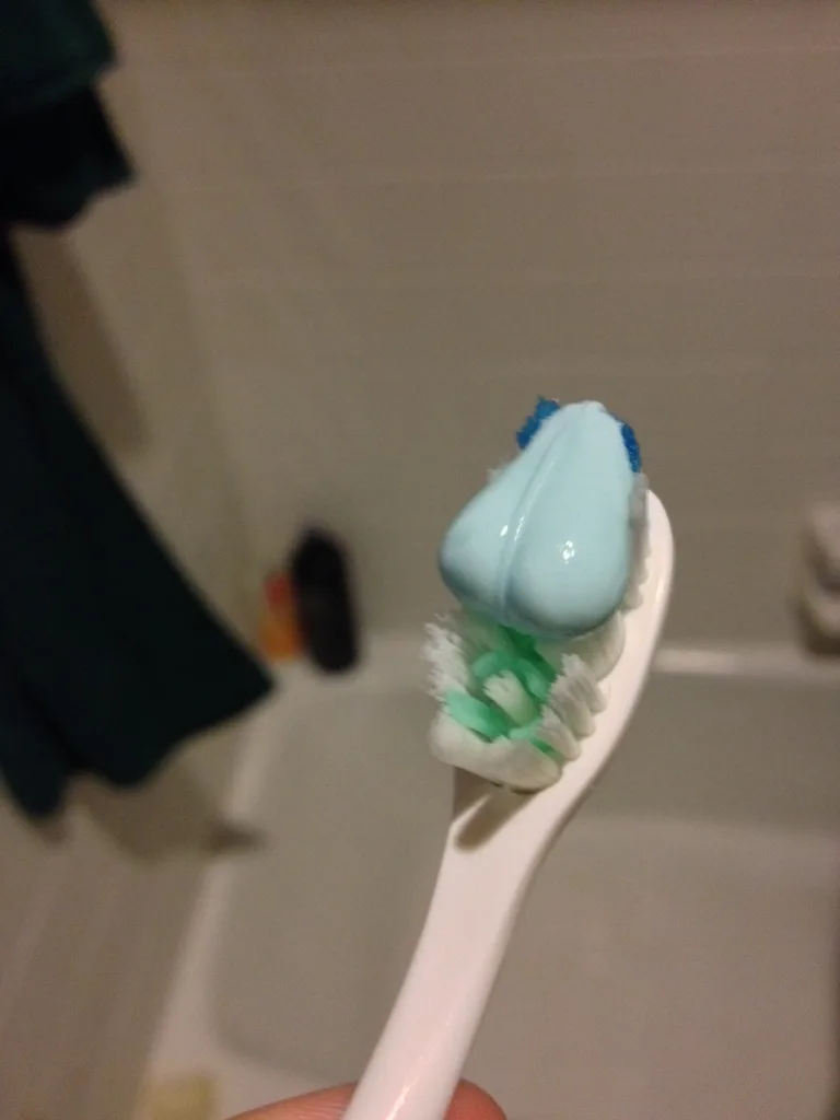 A close-up photo shows a white toothbrush with a large, oddly shaped dollop of light blue toothpaste on its bristles…