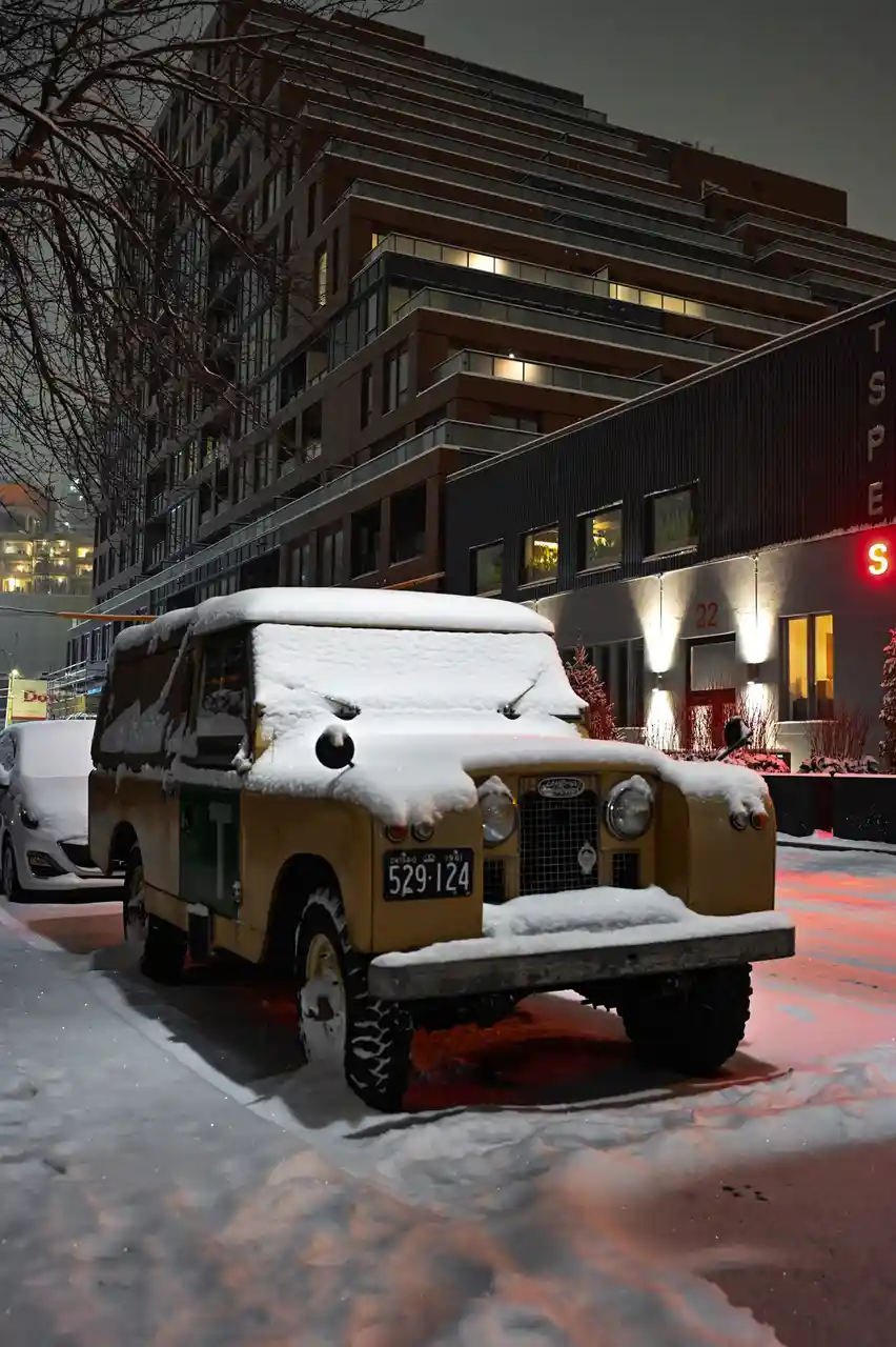 A vintage yellow Land Rover is parked on a snowy city street at night, completely covered in snow, with modern buildings and…