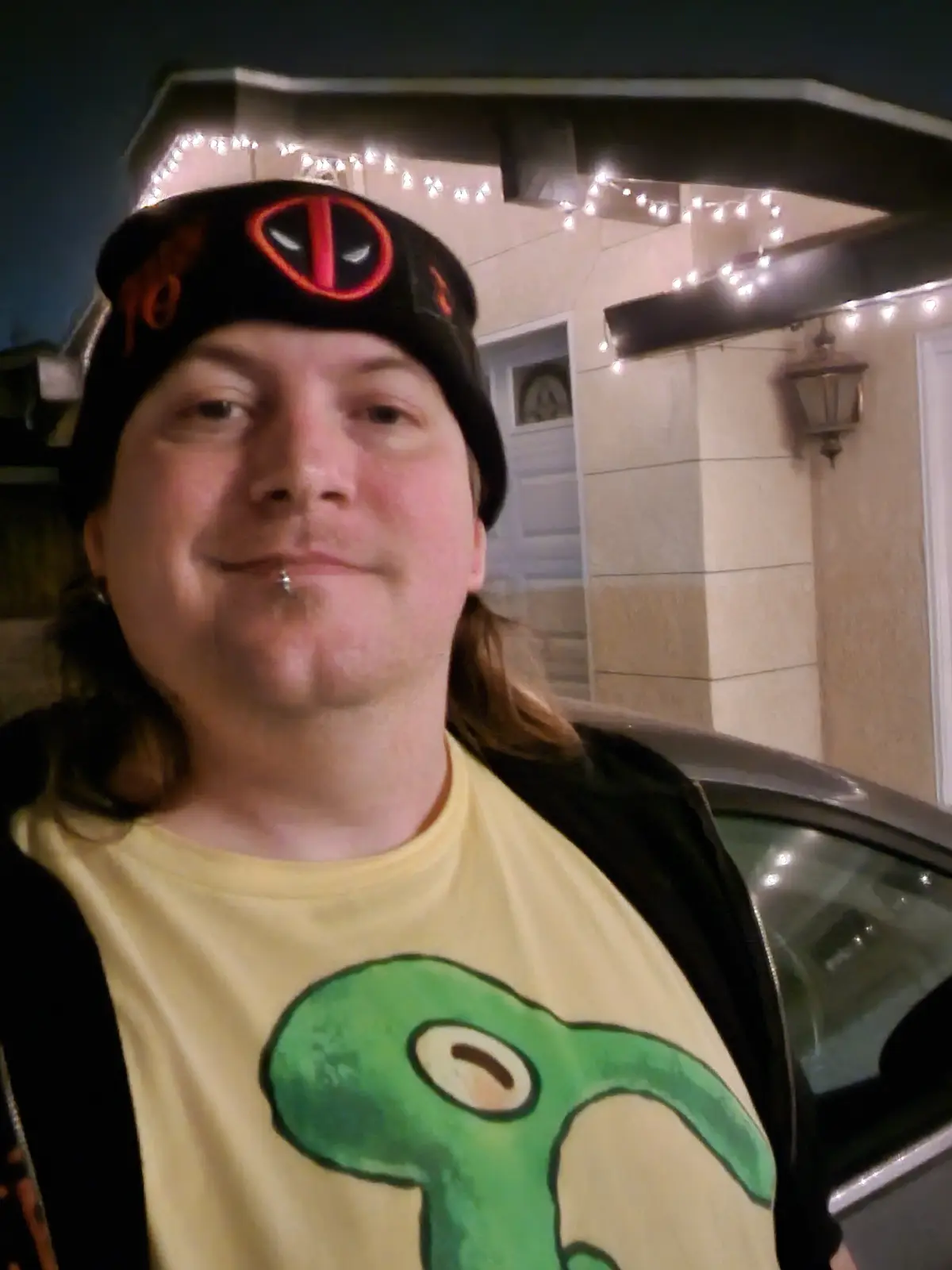 A person with long hair, a lip piercing, and a black beanie featuring the Deadpool logo stands outside at night in front of…