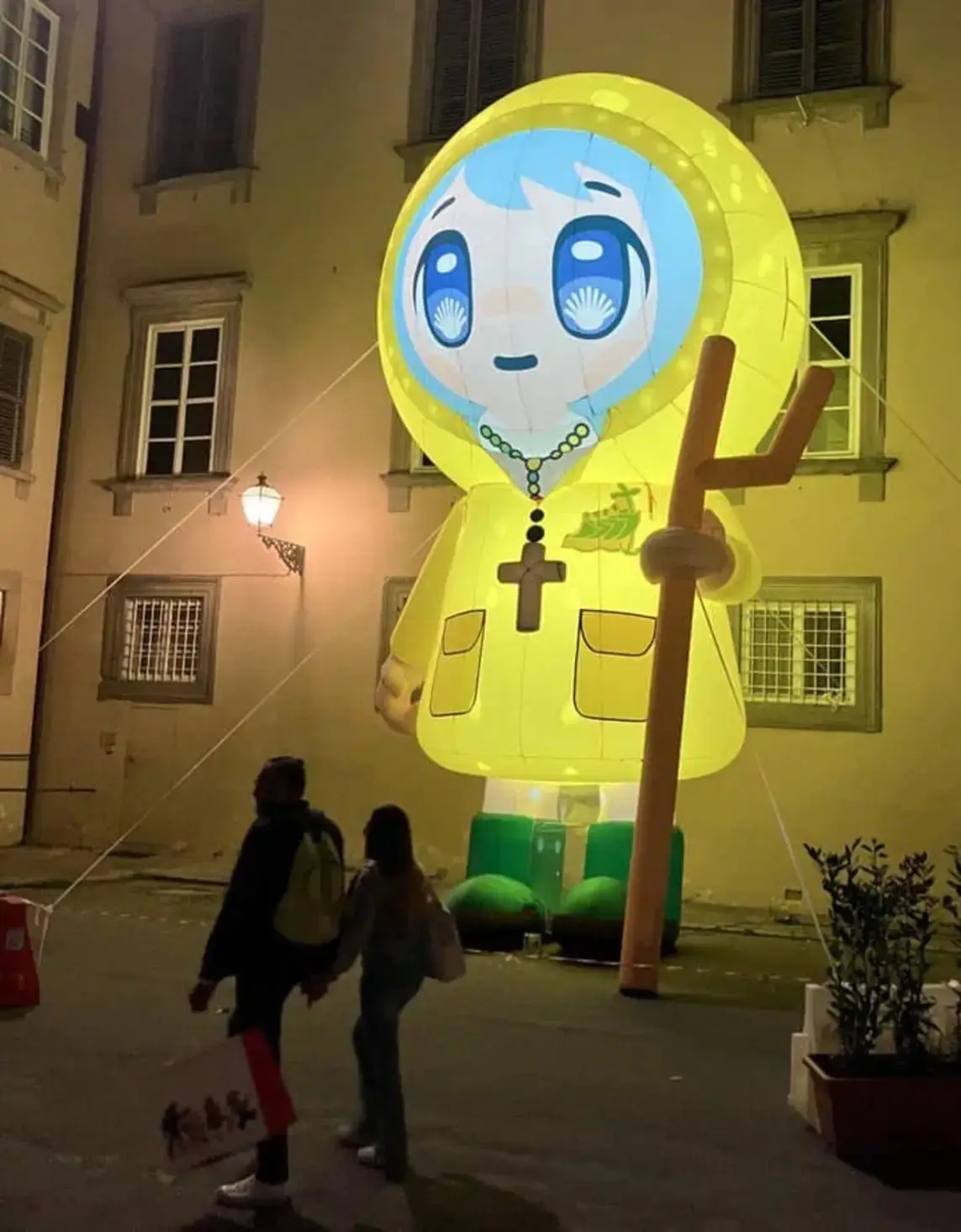 A large, illuminated yellow balloon shaped like a cartoon character with blue hair and eyes, wearing a yellow robe with a…