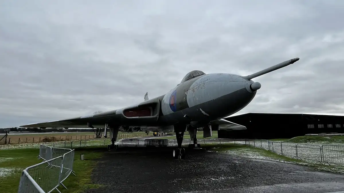 A weathered, grey military jet aircraft, likely a British WWII-era plane with a roundel insignia, is displayed outdoors on a…
