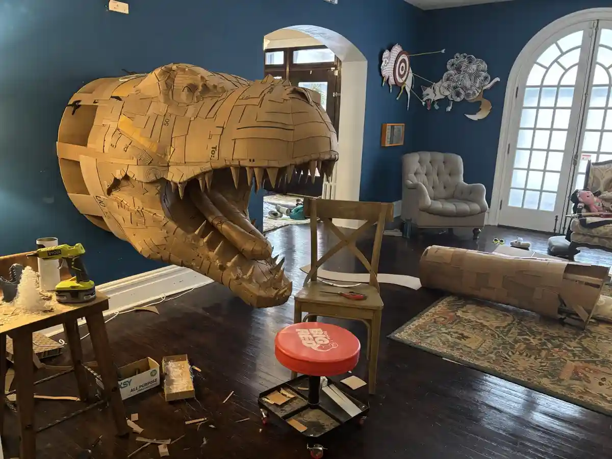 A large, intricate cardboard sculpture of a crocodile's head with its mouth open and sharp teeth, is positioned in a living…