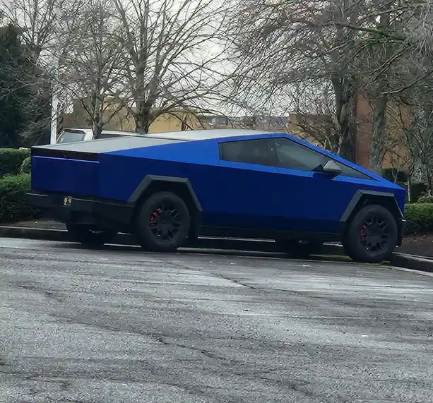 A bright blue Tesla Cybertruck is parked at an angle in a paved parking lot, surrounded by bare trees and shrubs under an…