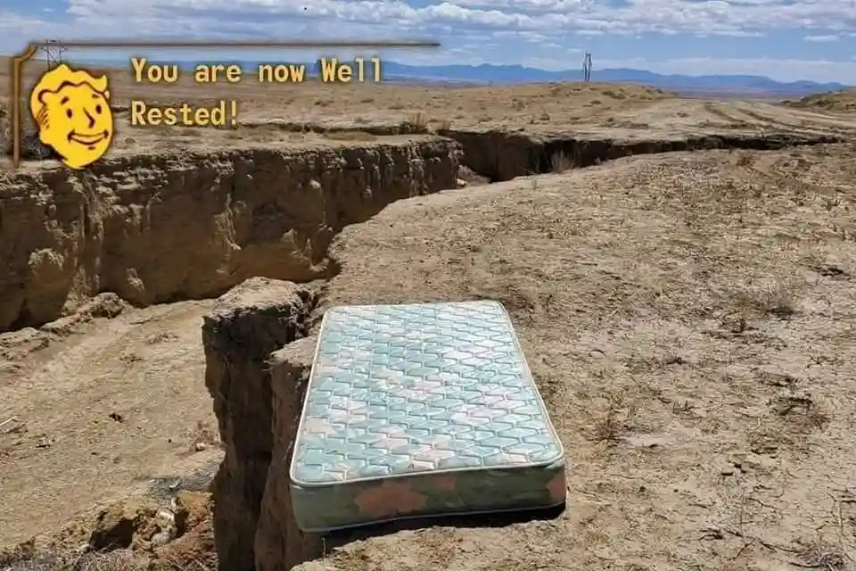 A mattress sits precariously on the edge of a deep, dry canyon in a barren desert landscape under a partly cloudy sky. The…