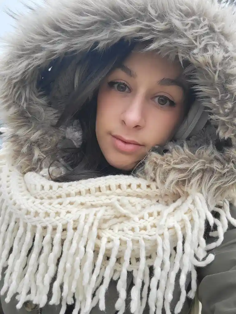 A person wearing a heavy winter coat with a large, fluffy grey fur-lined hood and a thick, cream-colored knitted scarf with…