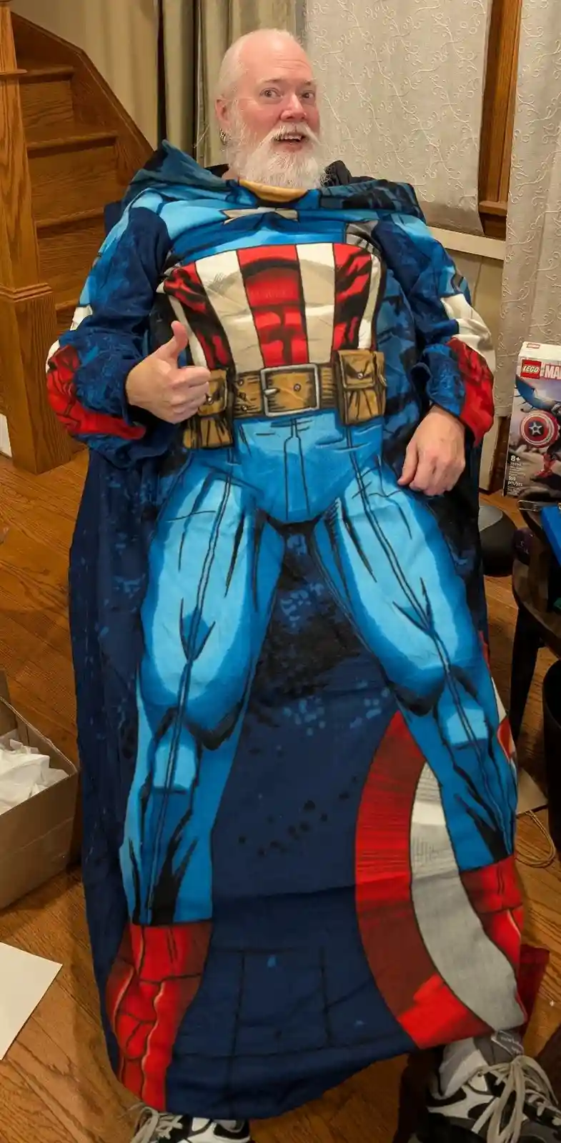 A man with a white beard and earring is wrapped in a large, full-body Captain America blanket, giving a thumbs-up while…