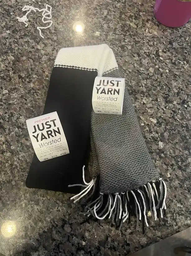 Two knitted scarves, one black and one gray with white fringe, lie on a speckled countertop, each with a 'PREMIER JUST YARN…