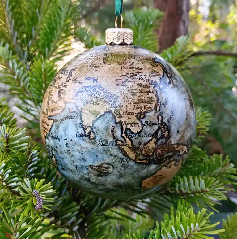 A globe-shaped Christmas ornament with a vintage map of Asia and the Indian Ocean hangs from a green pine tree branch. The…