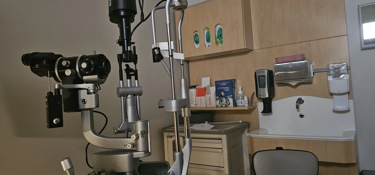 A photo of an eye examination room with a large, complex ophthalmology machine in the foreground, a sink, hand sanitizer…