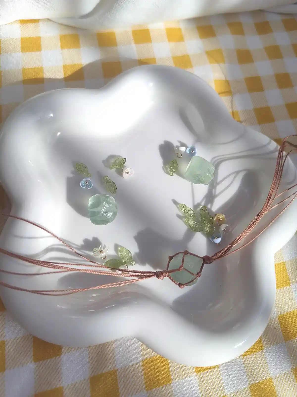 A white, cloud-shaped ceramic dish holds a handmade macrame necklace with a large, rough-cut sea glass pendant and smaller…