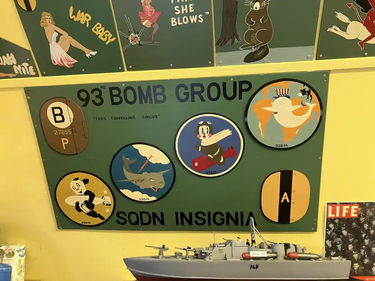 A green plaque labeled '93RD BOMB GROUP' displays various WWII-era squadron insignias, including cartoon animals like a…