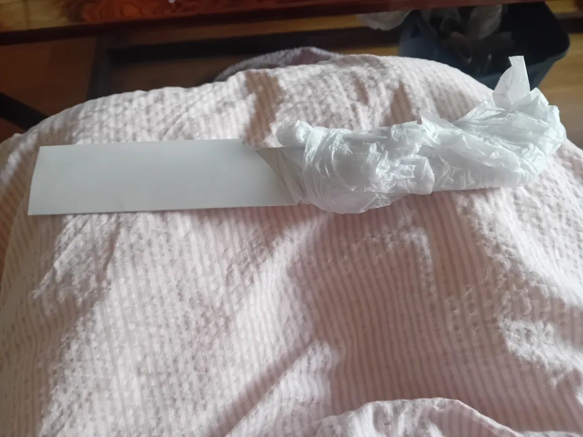 A crumpled white plastic bag sits on a pink and white striped bedspread, with a plain white rectangular piece of paper…
