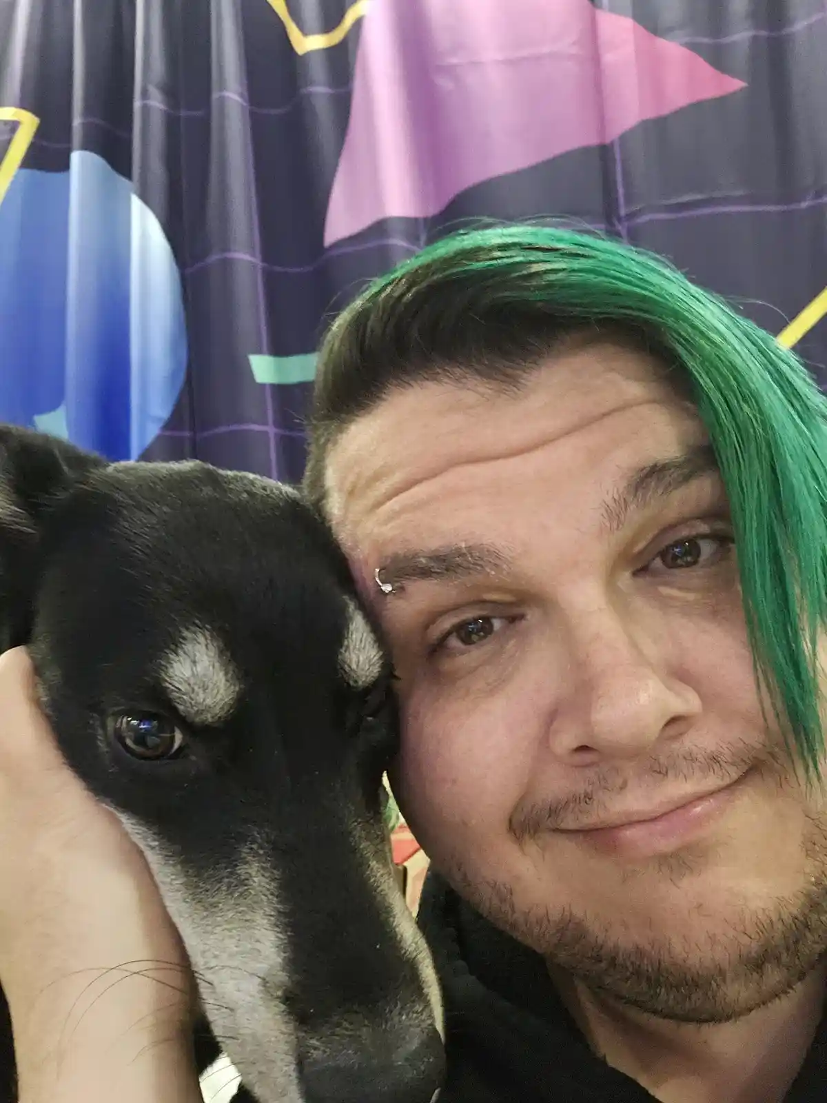 A selfie shows a man with green-dyed hair and a forehead piercing holding a black and white dog close to his face. The dog's…