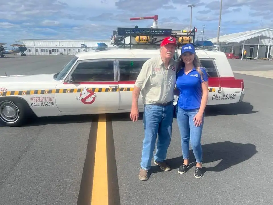 An older man and a younger woman stand smiling in front of a white hearse-style car decorated with Ghostbusters branding…