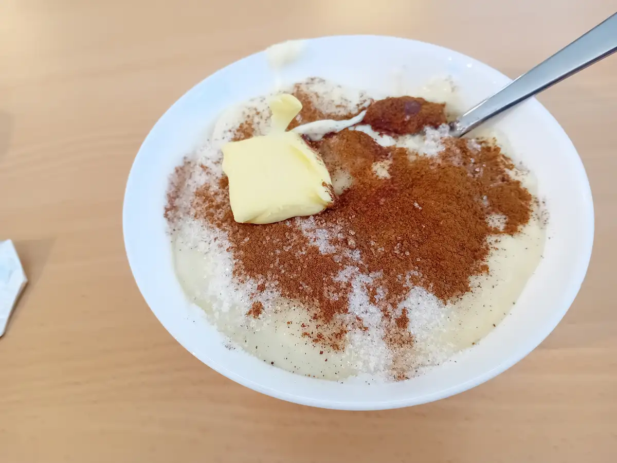 A white bowl on a wooden table contains a creamy dessert topped with a pat of butter, granulated sugar, and brown powder…