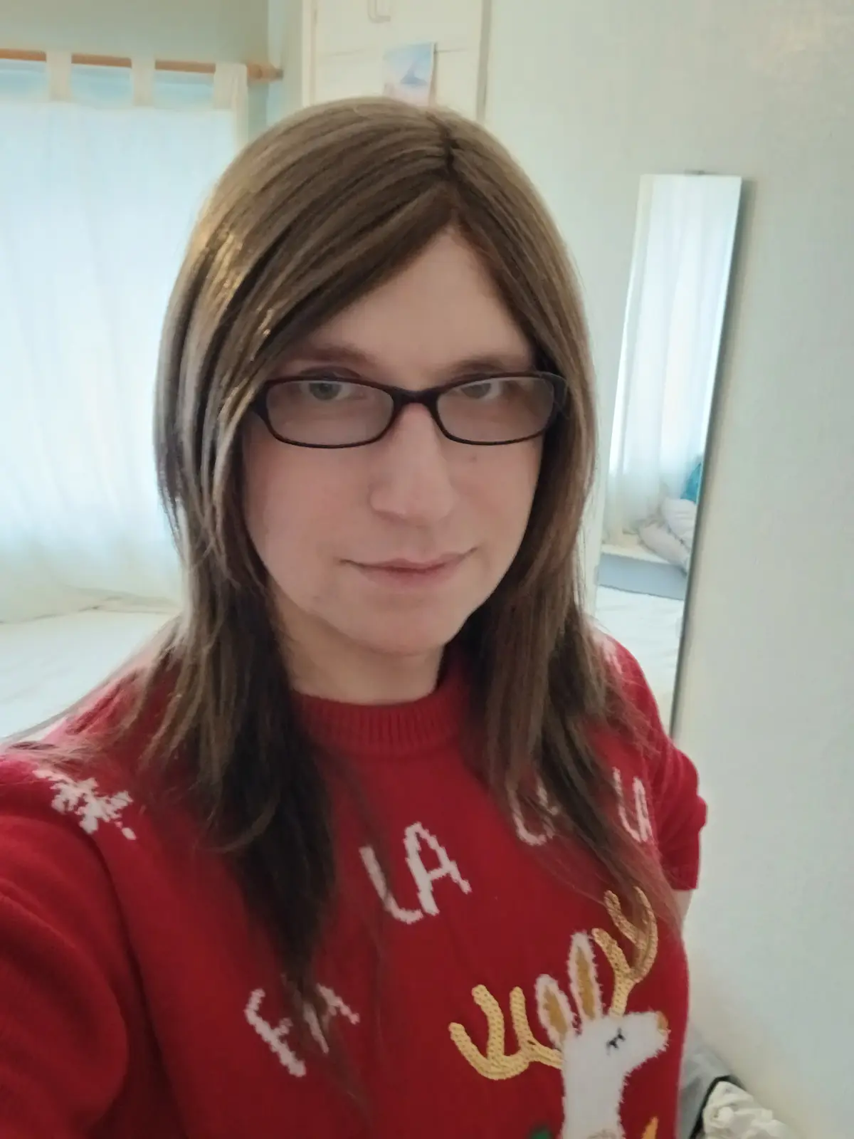 A person with long brown hair and glasses wears a red Christmas sweater featuring a white deer with antlers and the text 'la…