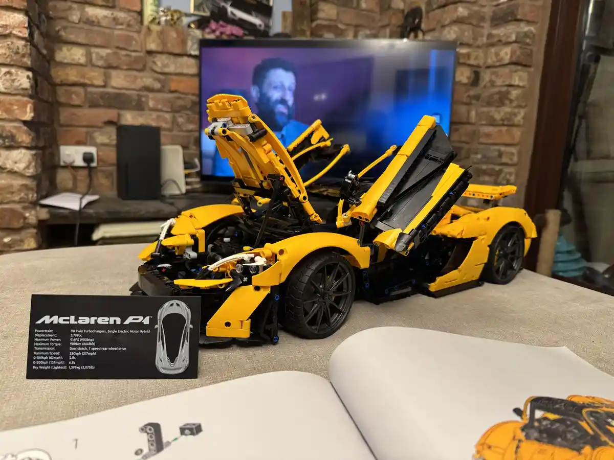 A yellow LEGO McLaren P1 model with its doors and hood open sits on a table, next to its instruction manual and a card…