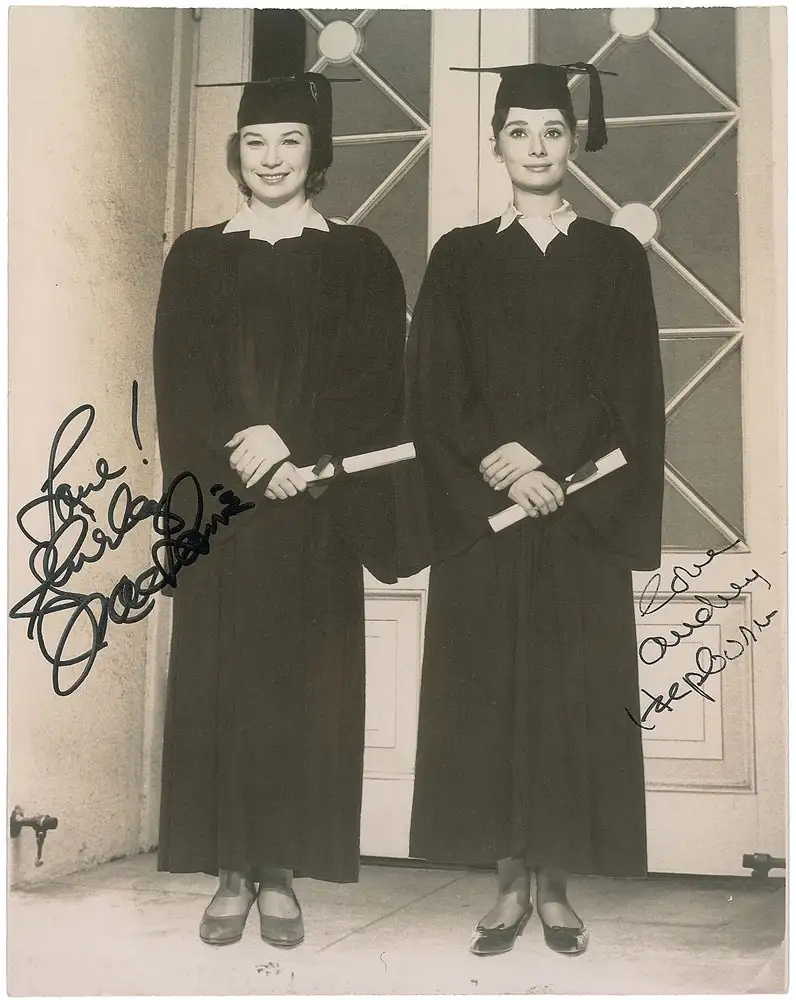 A black-and-white photo shows two women in graduation gowns and caps, each holding a diploma. The woman on the left has a…
