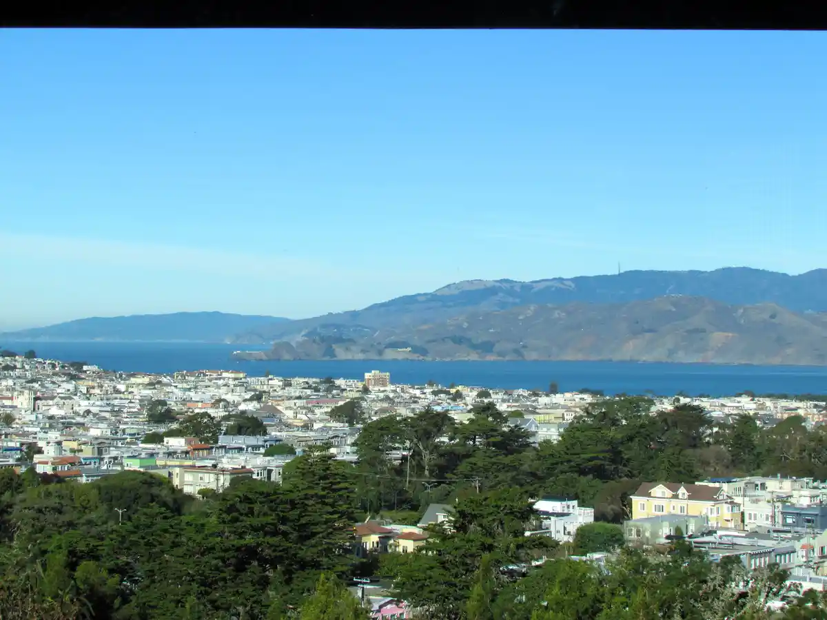 A scenic view of a coastal city with colorful houses, green trees, and distant mountains under a clear blue sky, overlaid…