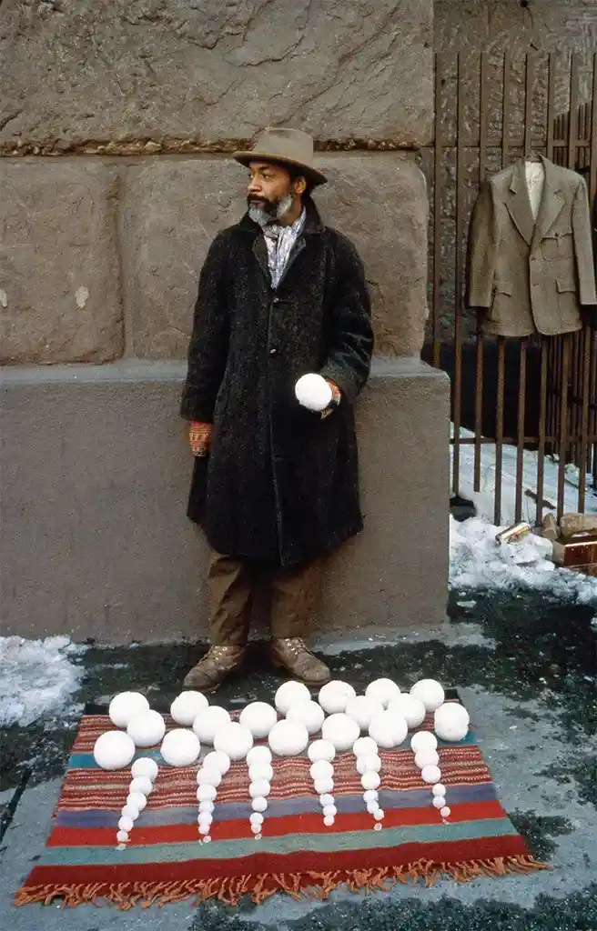 A man in a dark coat and hat stands outdoors on a snowy day, holding a snowball. In front of him, arranged on a colorful…