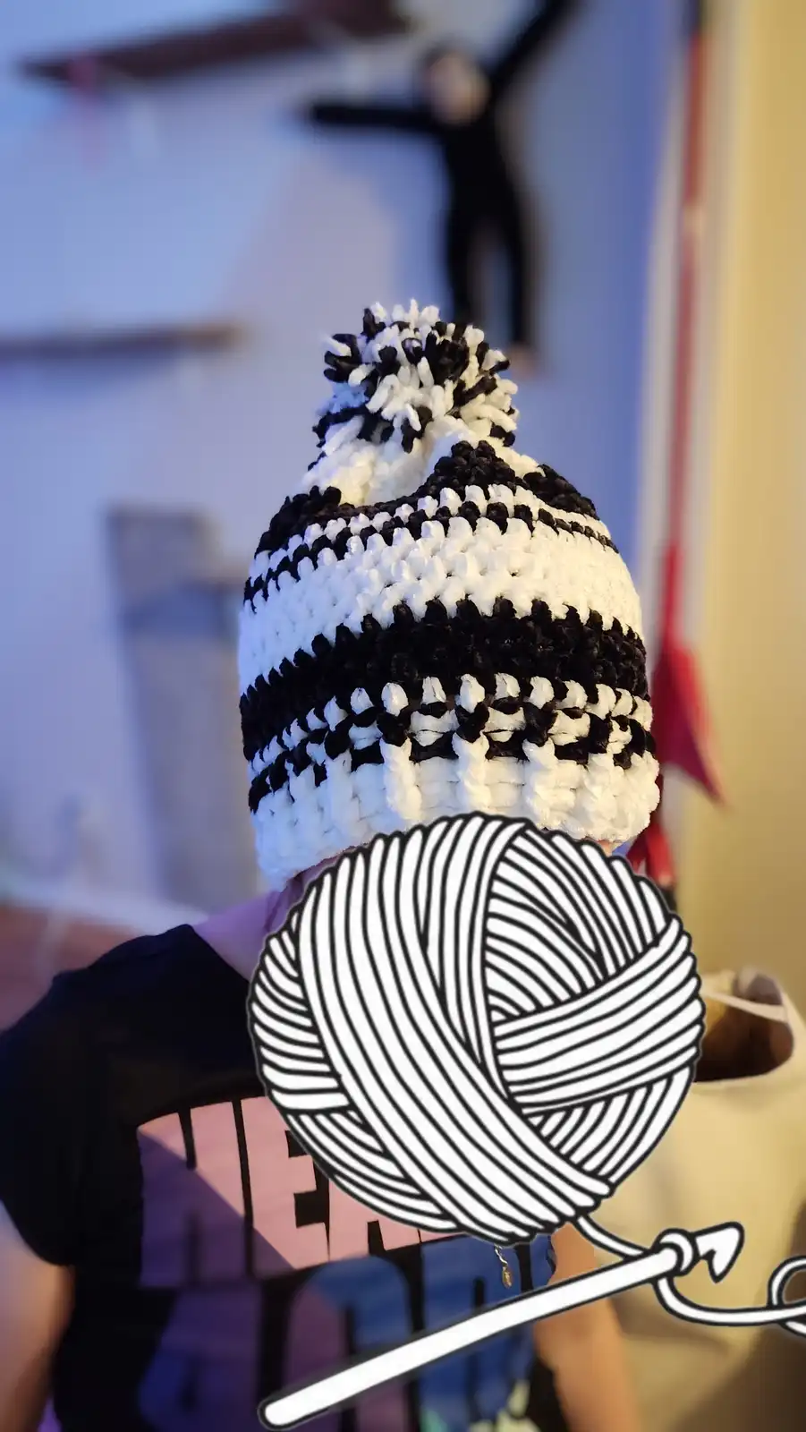 A person viewed from behind wears a black and white striped crocheted beanie with a pom-pom on top. A large cartoon…