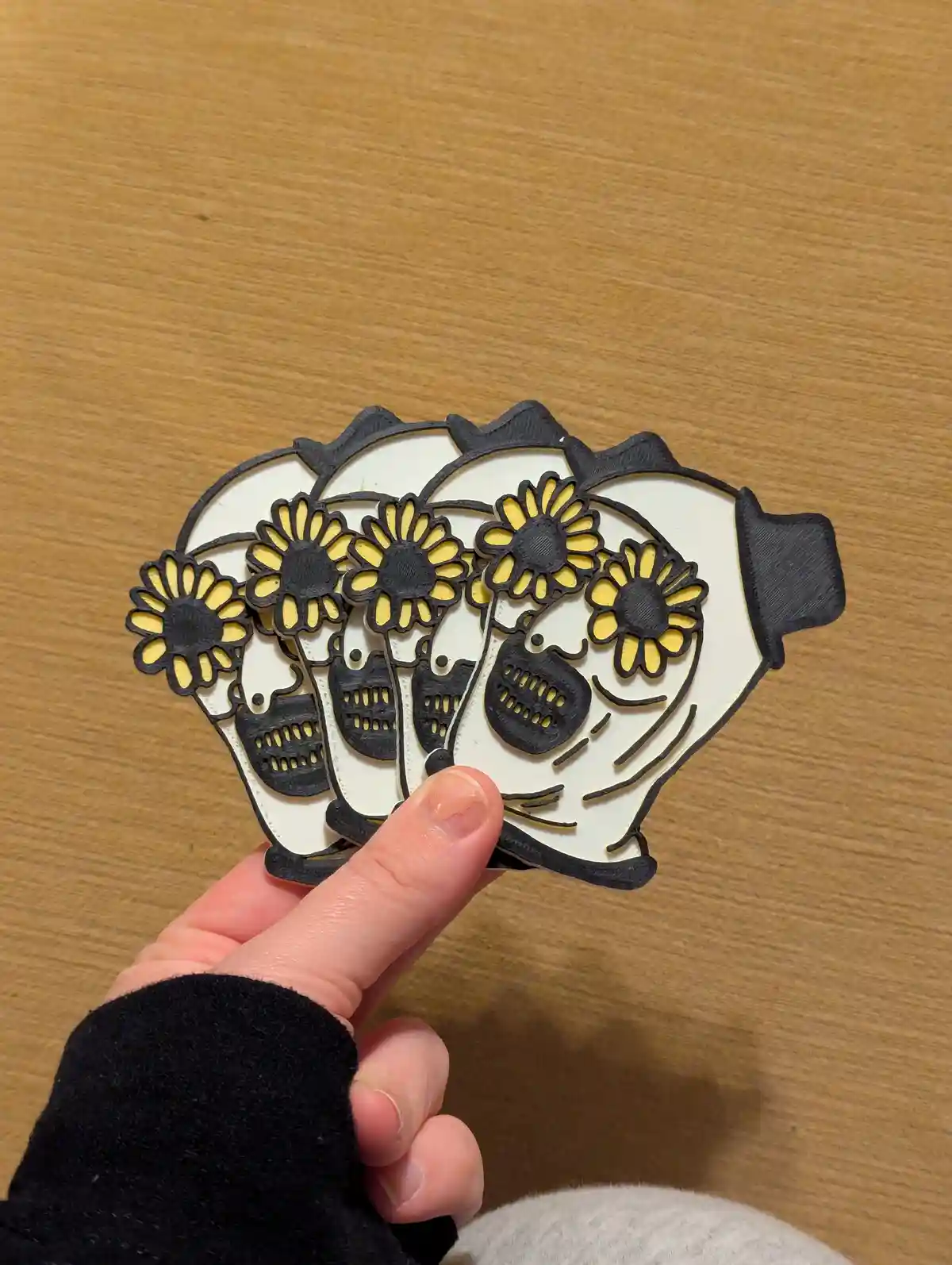 A hand holds three 3D-printed, white and black sugar skull-shaped coasters, each decorated with yellow sunflowers, against a…