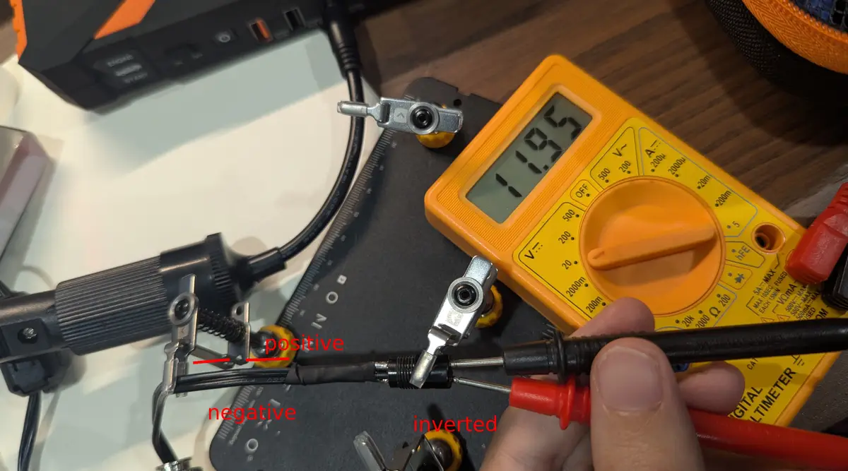 A close-up photo shows a hand holding a red multimeter probe connected to a black wire, with a yellow digital multimeter…