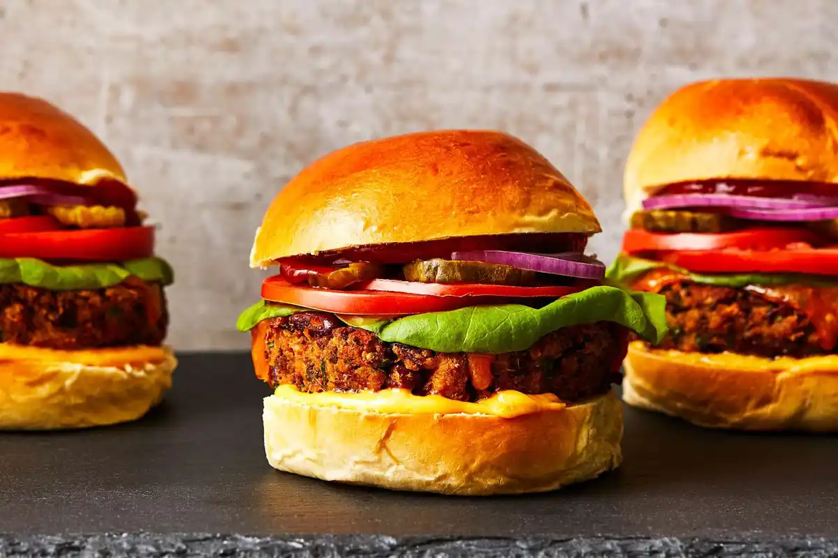 Three appetizing veggie burgers with golden buns, lettuce, tomato, onion, and pickles are arranged on a dark slate surface…