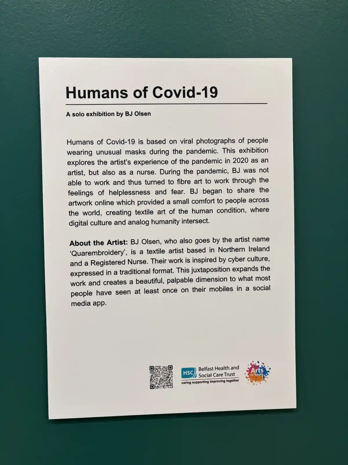 A white informational plaque mounted on a dark green wall describes an art exhibition titled 'Humans of Covid-19' by artist…