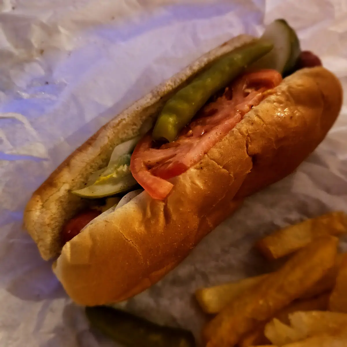 A close-up photo of a hot dog in a bun with visible toppings including a pickle spear, tomato slice, and possibly onions…