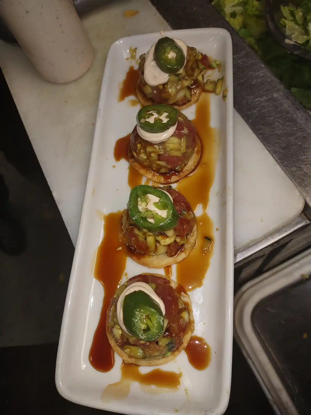 a white rectangular plate holds four small, round appetizers topped with raw meat, chopped vegetables, a dollop of white…