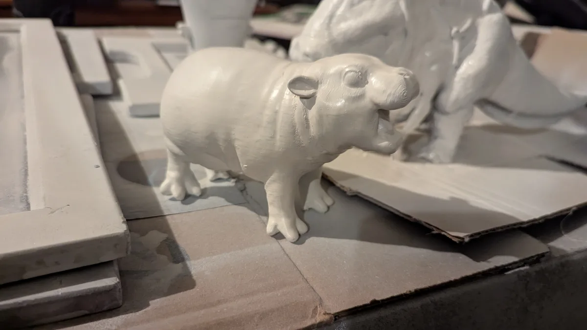 A white ceramic figurine of a hippopotamus with its mouth wide open, standing on cardboard amidst other white ceramic…