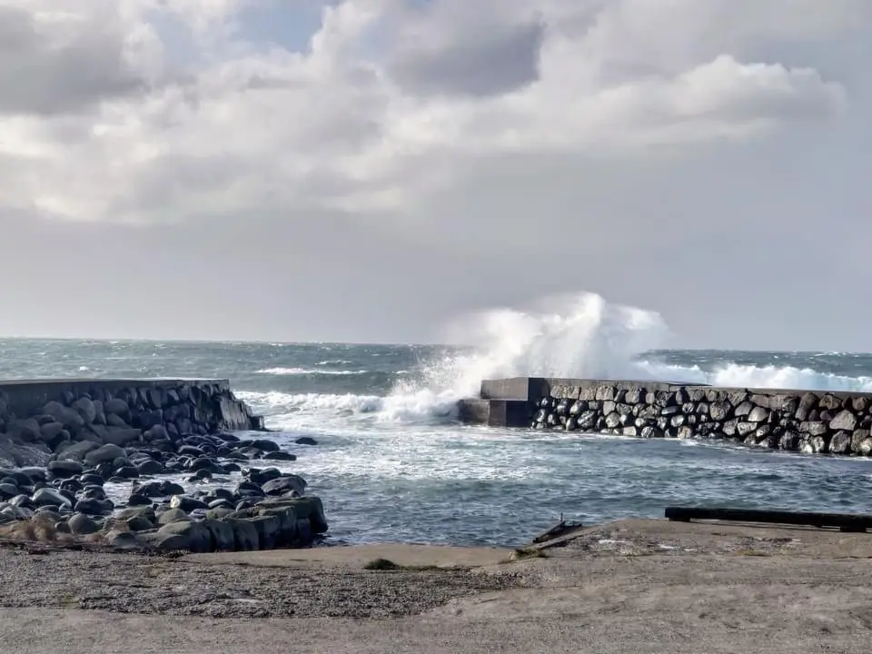 A powerful ocean wave crashes violently against a concrete and rock breakwater under a cloudy sky. The overlaid text reads…
