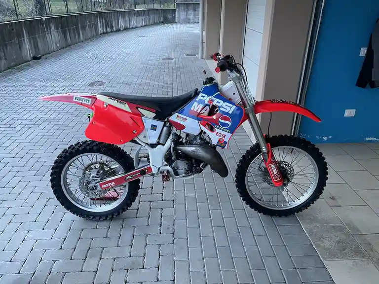 A red and white Honda dirt bike is parked on a gray brick patio, featuring prominent Pepsi branding on its side panel. The…