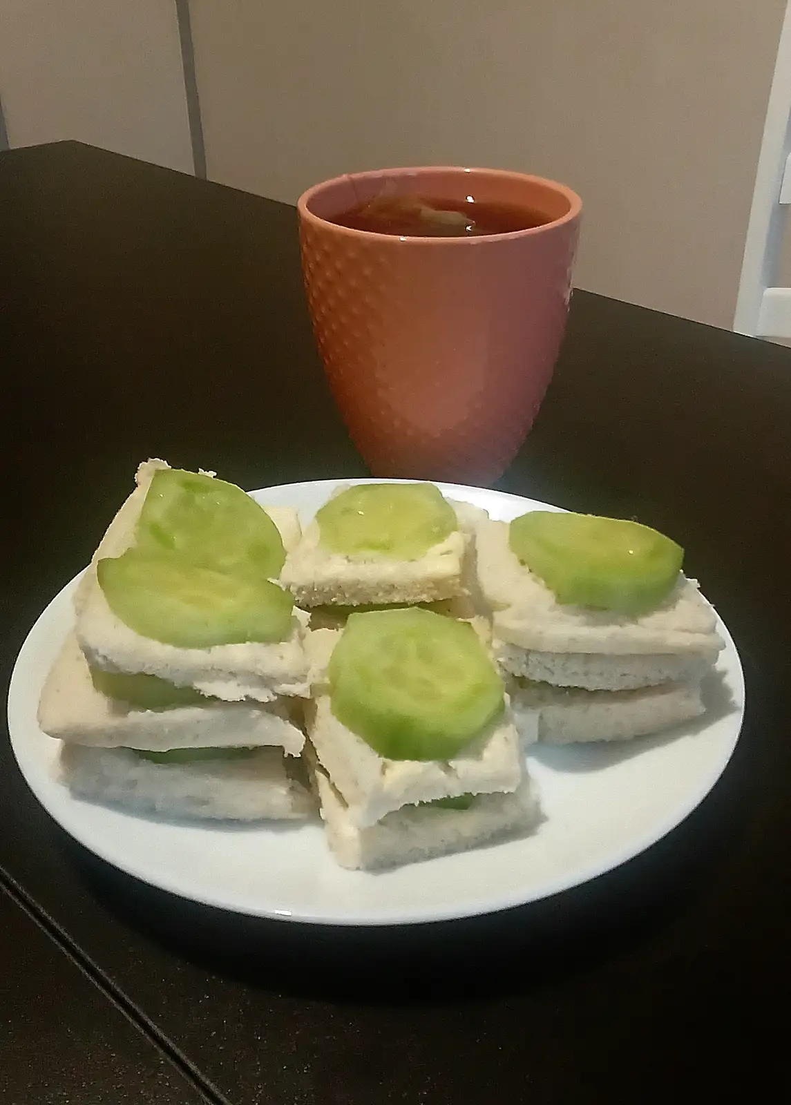 A white plate holds several small sandwiches made with white bread and topped with green cucumber slices, placed on a dark…