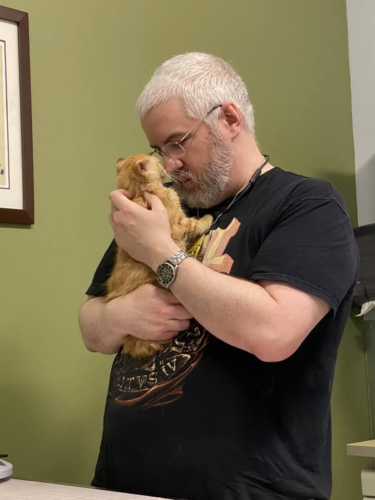A man with white hair and glasses is holding a small, fluffy orange kitten close to his face, appearing to nuzzle or kiss…