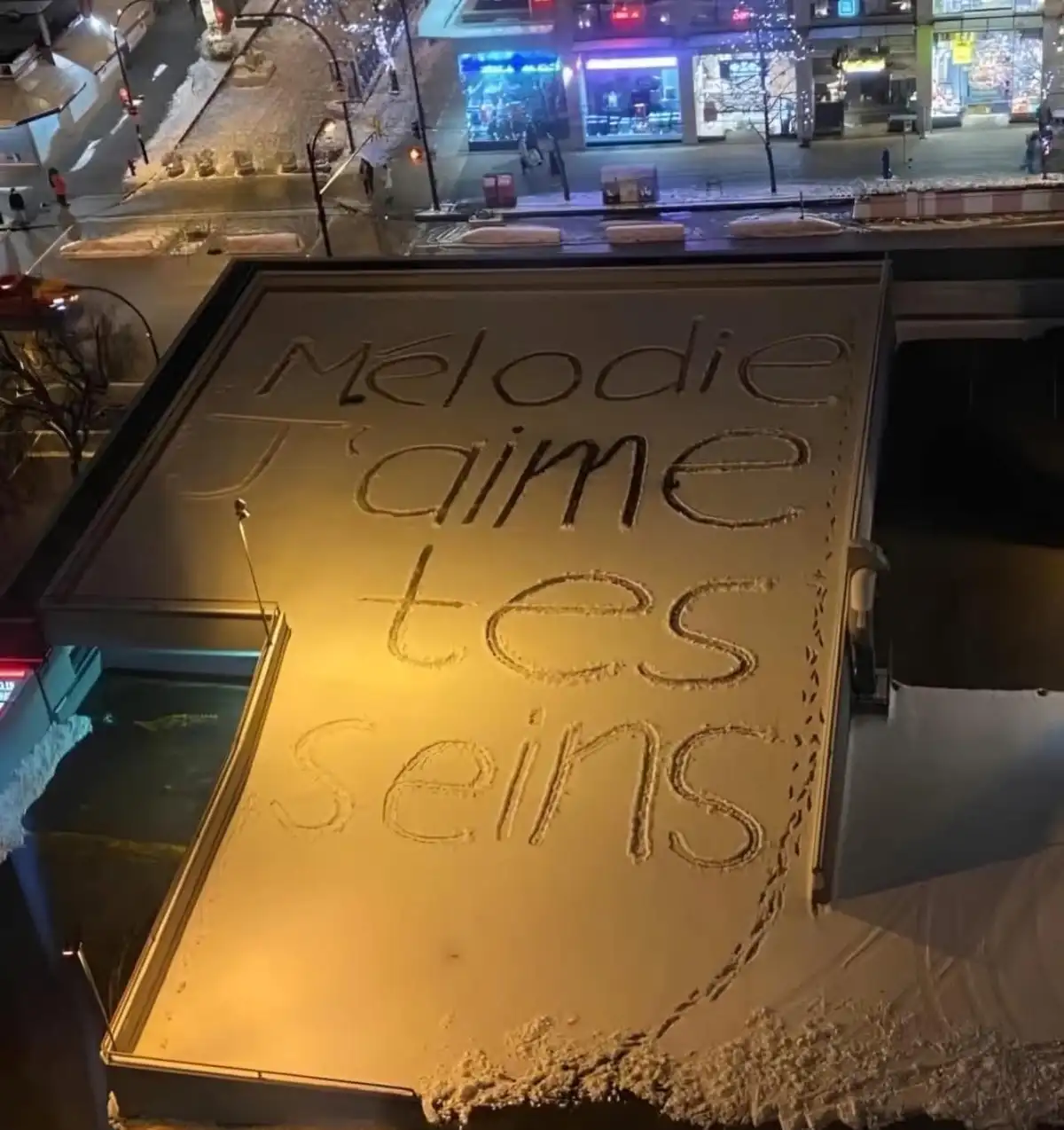 A snowy rooftop at night has the French phrase 'Mélodie J'aime tes seins' written in snow using a finger or stick. The text…