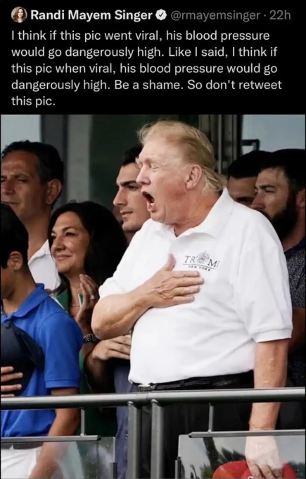 A man in a white polo shirt with 'TRUMP NEW YORK' on the chest stands with his hand over his heart, mouth open as if…