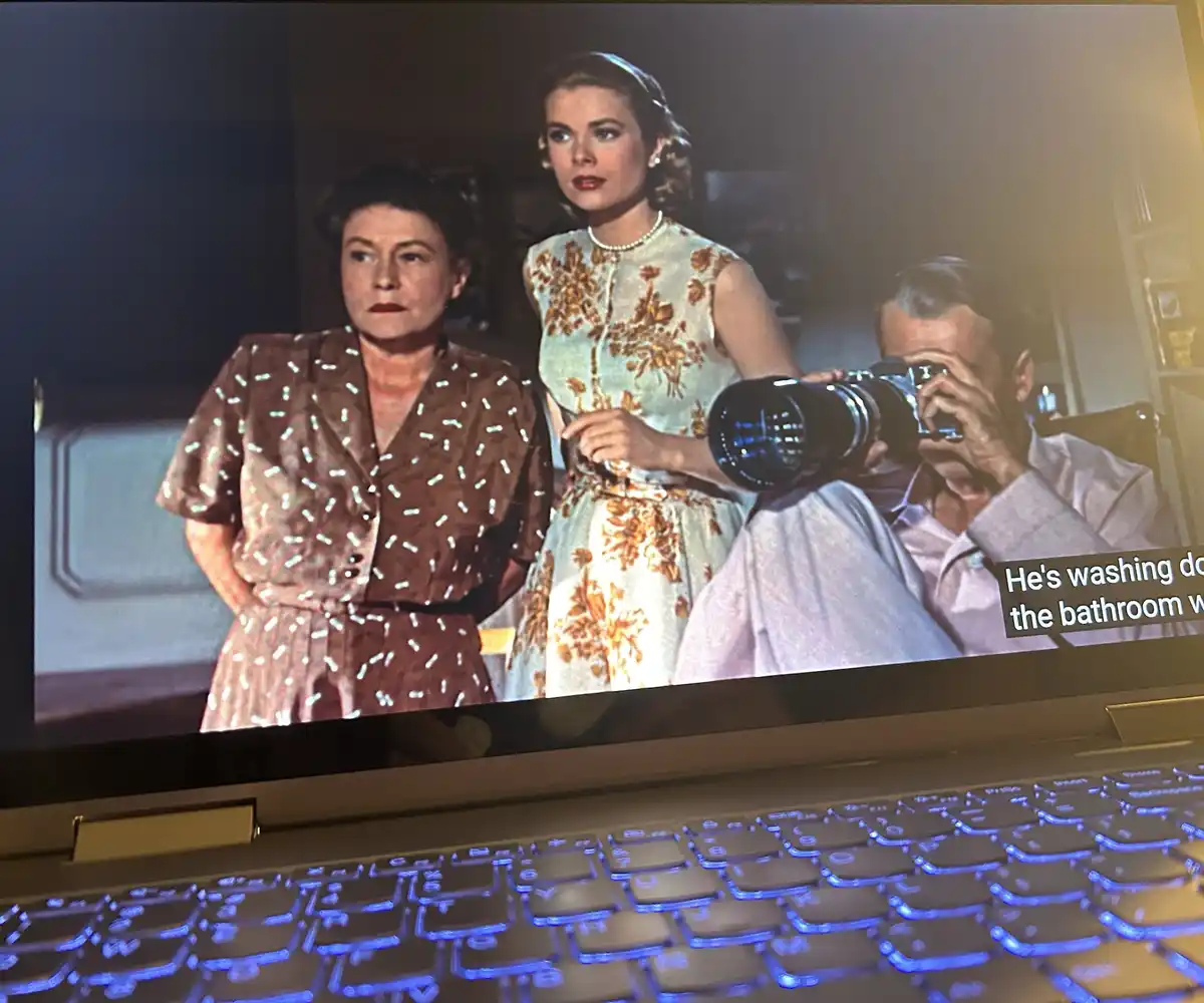 A laptop screen displays a scene from a vintage film featuring two women and a man holding a large camera. The man is…
