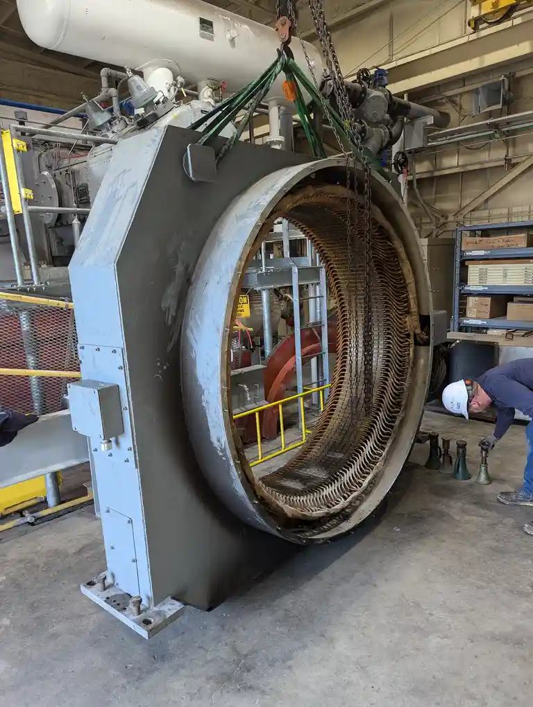 A large industrial machine, possibly a generator rotor, is suspended by chains in a factory setting. A worker in a hard hat…