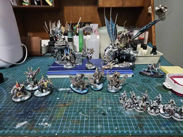 A cluttered hobbyist's workspace shows numerous intricately painted fantasy miniatures arranged on a green cutting mat with…