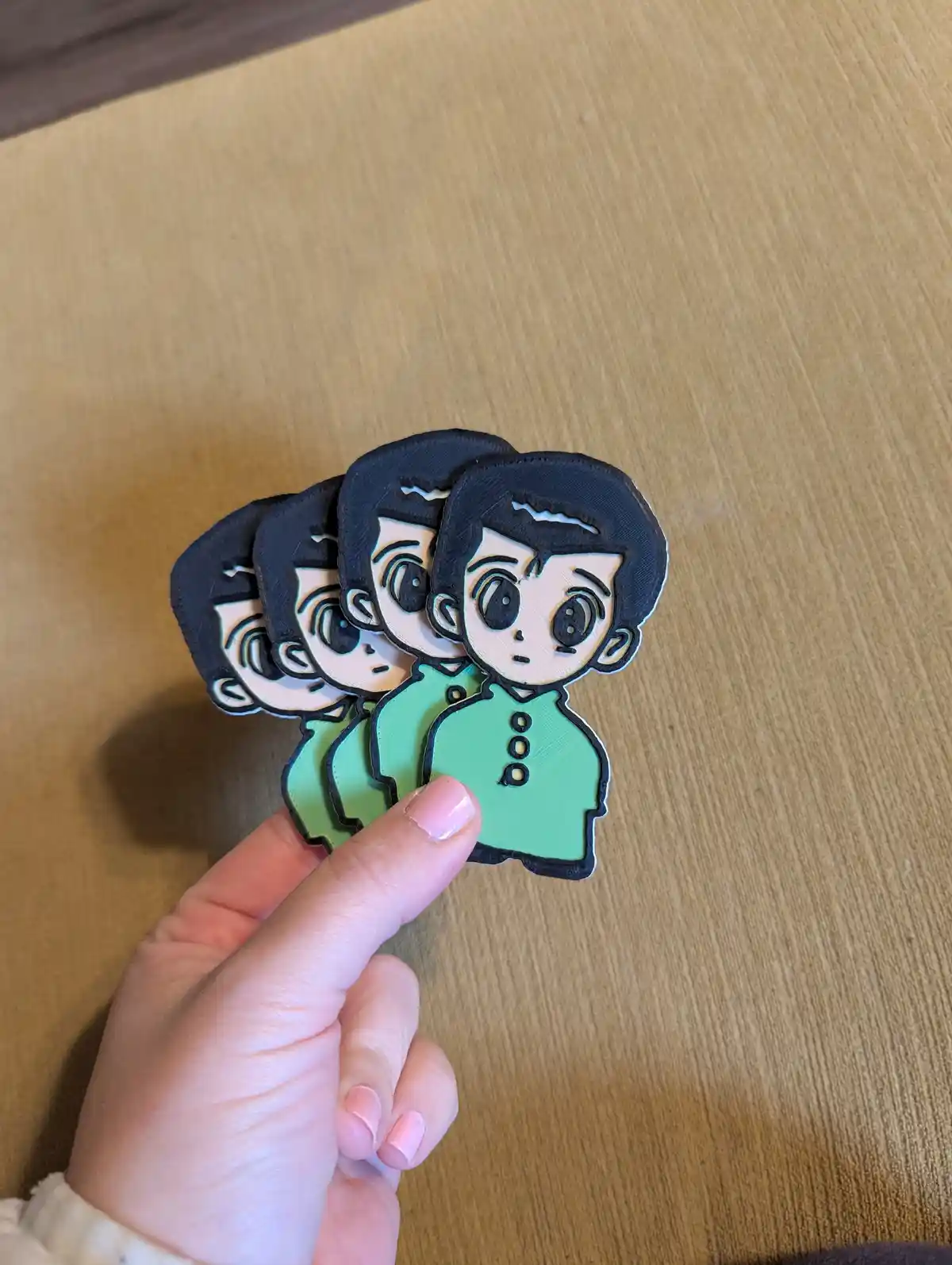 A hand holds a fanned-out stack of five identical 3D-printed anime-style character cutouts, each depicting a boy with black…