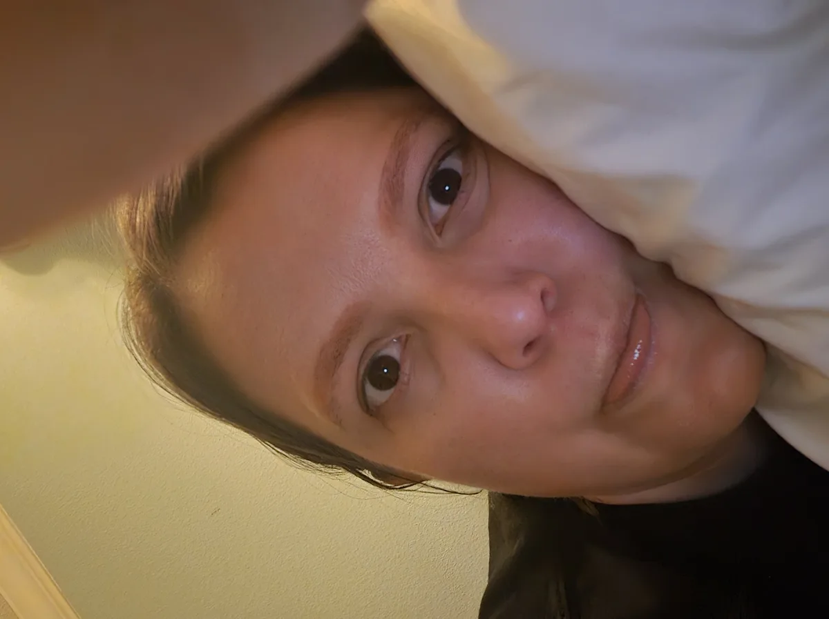 A person with brown eyes and light brown hair looks directly at the camera with a slight smile while holding a white pillow…