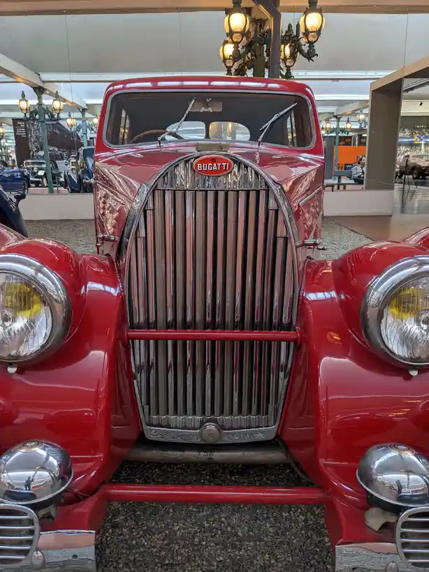 A close-up frontal view of a shiny red vintage Bugatti automobile, showcasing its iconic chrome grille, round headlights…