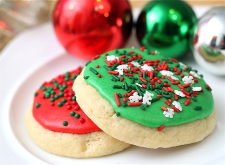 two round sugar cookies with red and green icing and festive sprinkles sit on a white plate, with shiny red, green, and…