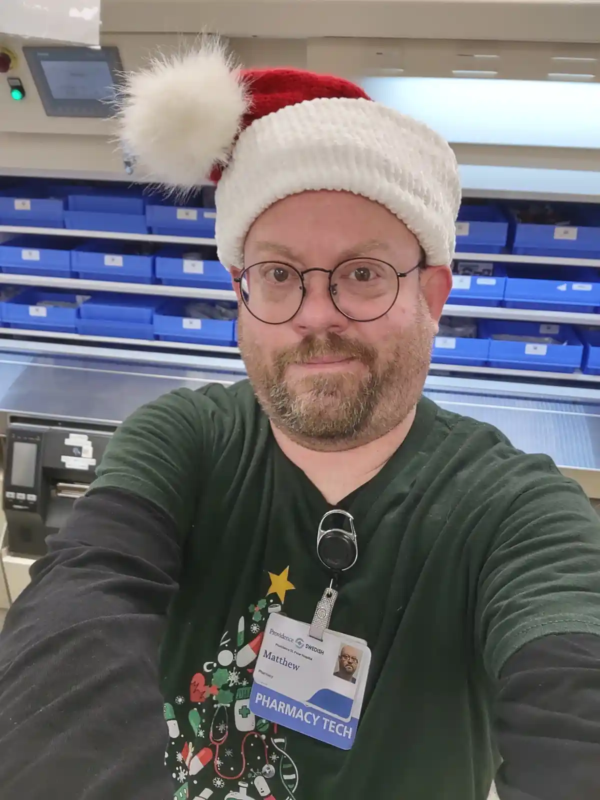 A man wearing a red and white Santa hat and a green holiday-themed t-shirt with medical symbols, takes a selfie in a…