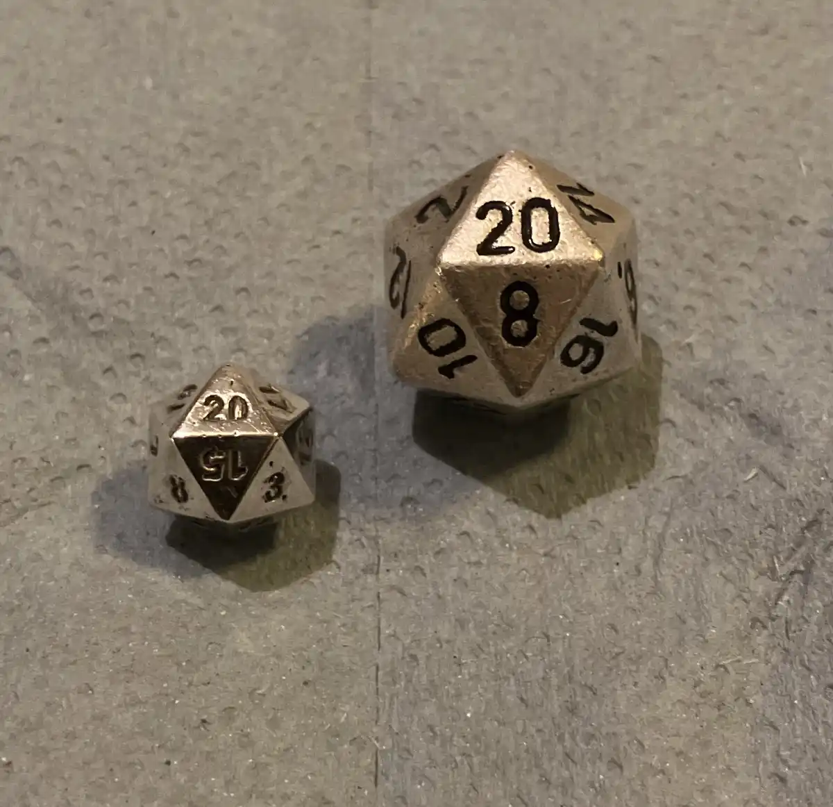 Two metallic 20-sided dice rest on a textured gray surface; the larger die shows '20' and '8', while the smaller die shows…