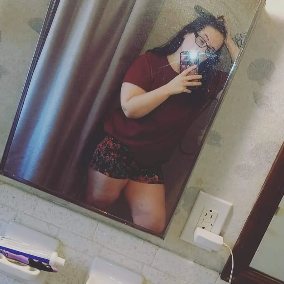 A person with glasses takes a mirror selfie in a bathroom, holding their phone with one hand while resting their other hand…